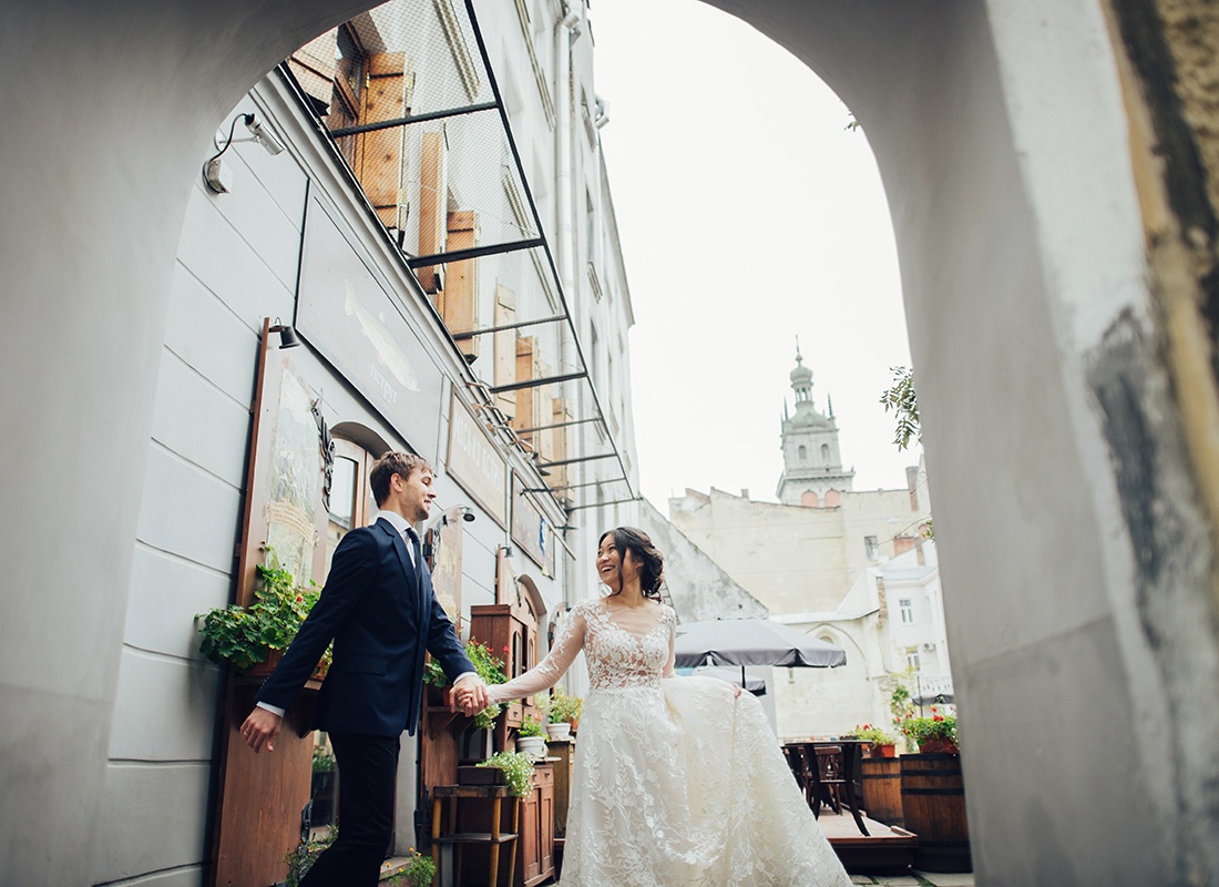 Foreign Wedding Insurance Quote - Portrait of a Cheerful Young Bride and Groom Holding Hands as They Walk Down an Alley in a Historic European City