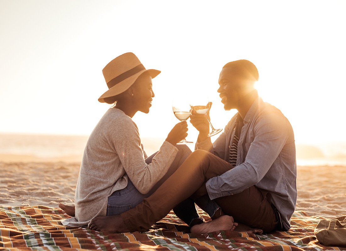 Personal Insurance - Portrait of a Young Couple Sitting on a Blanket on the Beach Sand at Sunset While Looking at Each Other and Enjoying a Glass of Wine