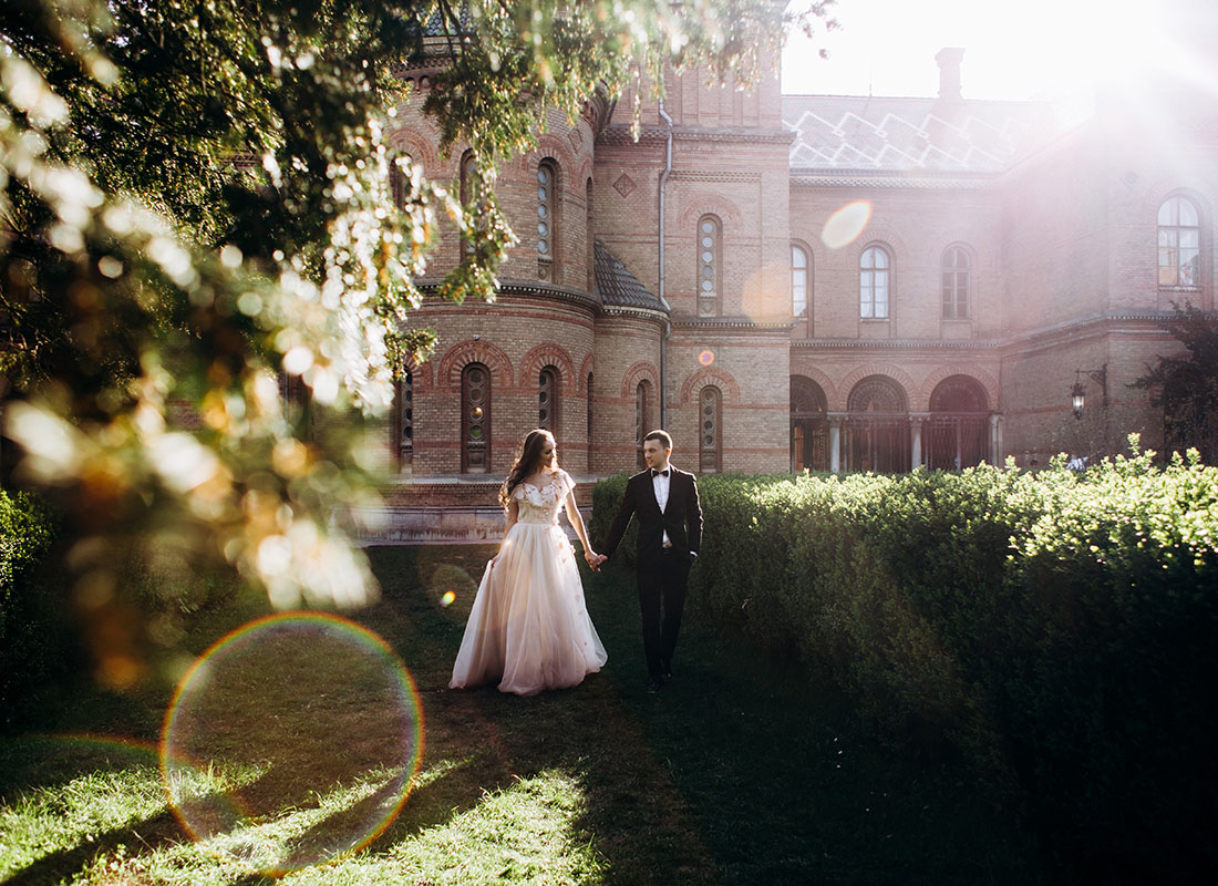 Wedding Insurance - Portrait of a Bride and Groom Walking on the Grass in the Sunlight with a Historic Mansion in the Background